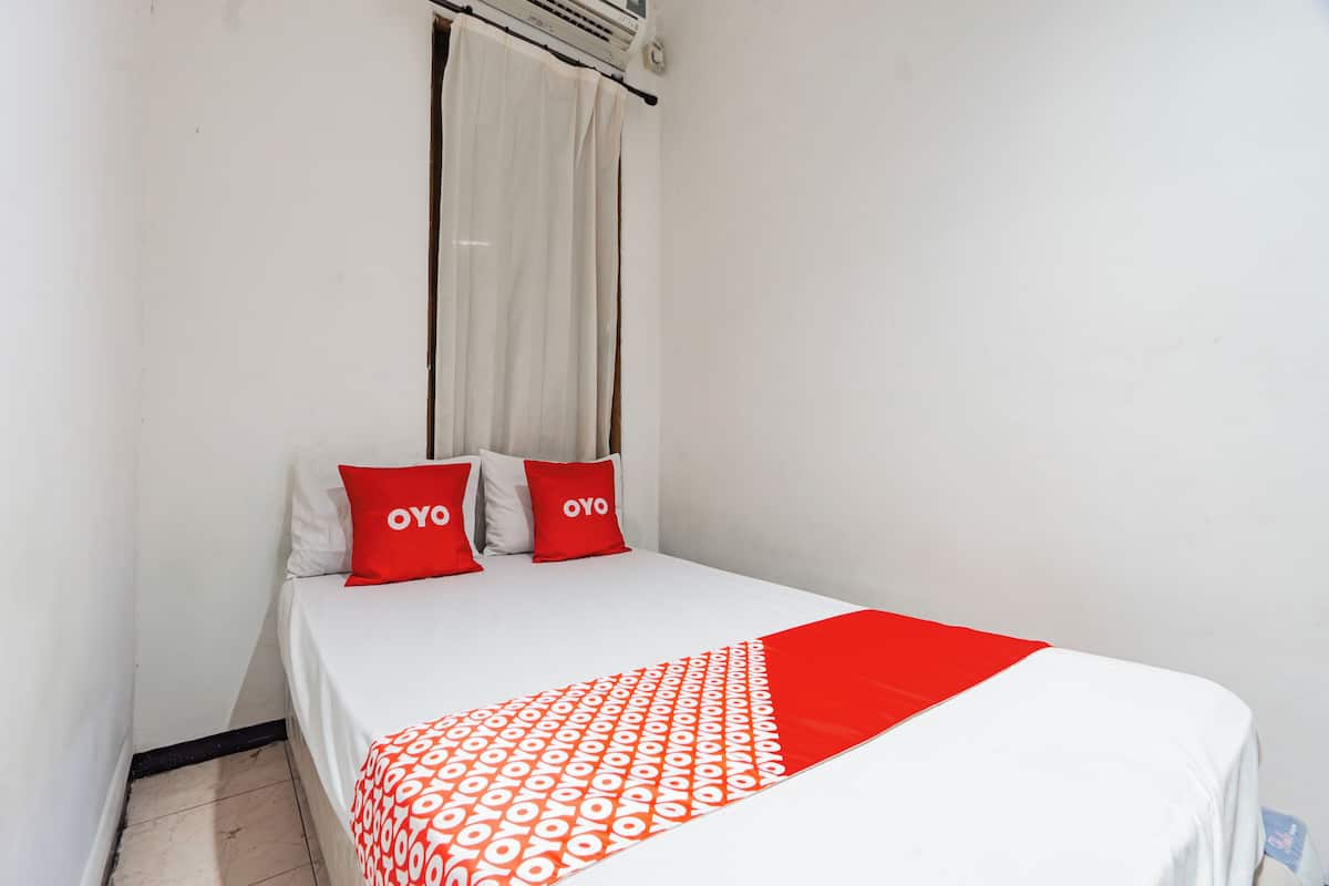 A single bed with a white and red bedspread, two red pillows with the word "OYO" on them, and a window with a white curtain.