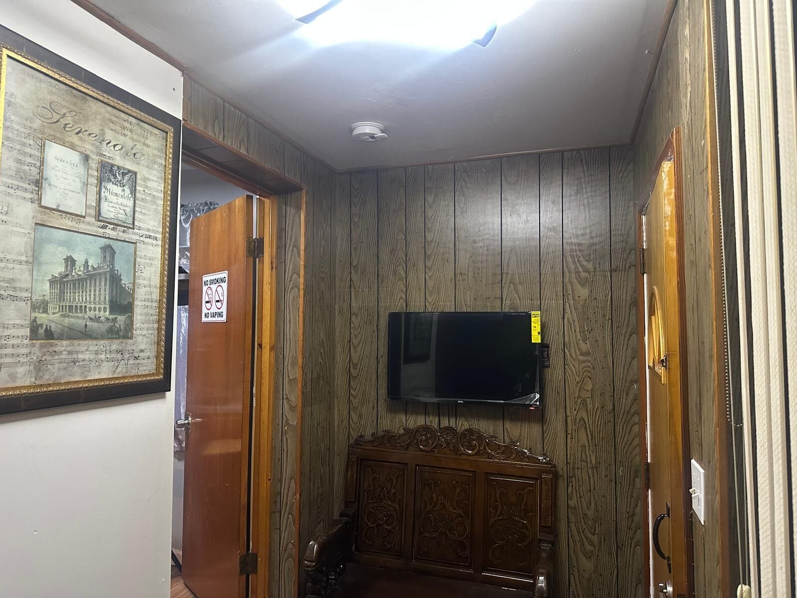 A room with wooden paneling, a mounted flat-screen TV, a framed picture on the wall, and a door with a 'No Smoking' sign.