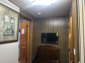 Una habitación con paneles de madera, un televisor de pantalla plana montado, un cuadro enmarcado en la pared y una puerta con un letrero de 'No Fumar'.