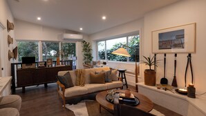 A modern living room with a sofa, coffee table, and a dining area. There is a large window with a view of trees, a lamp, and a framed picture on the wall. - Birds Nest of Topanga - Boho Chic and Minutes from Beach and Topanga Village!! (Topanga)