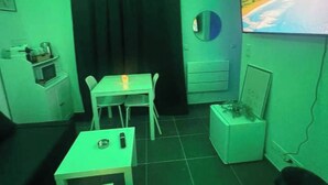 Ein Zimmer mit Fernseher, Tisch, Stühlen, einem kleinen Schrank und einem Vorhang.
