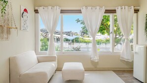 Una habitación con un sofá blanco, un pequeño reposapiés y una ventana con cortinas blancas.