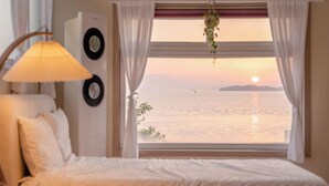 Un dormitorio con cama, lámpara, reloj en la pared y ventana con vista al mar al atardecer.