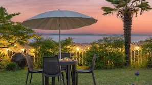 Un'area pranzo all'aperto con tavolo e sedie, un ombrellone e vista sul mare al tramonto.