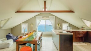 Un dormitorio cálido y luminoso en el ático con un gran ventanal con vistas al mar, una pequeña kitchenette y una mesa llena de color.