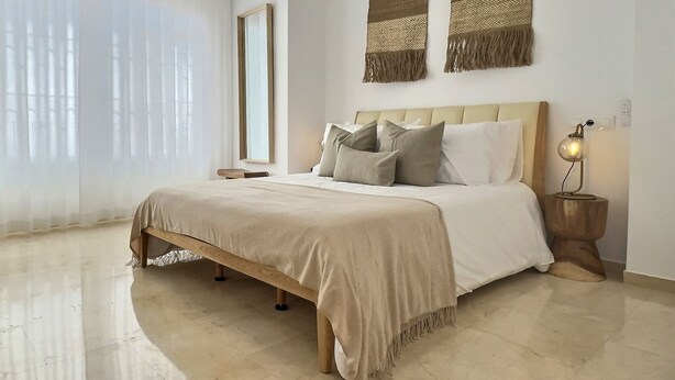Un dormitorio moderno con una cama grande, una mesita de noche, una lámpara y un amplio ventanal con cortinas translúcidas.