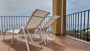 Un balcón con dos sillones blancos y piso de baldosas, con vistas a la costa, edificios y zonas verdes.