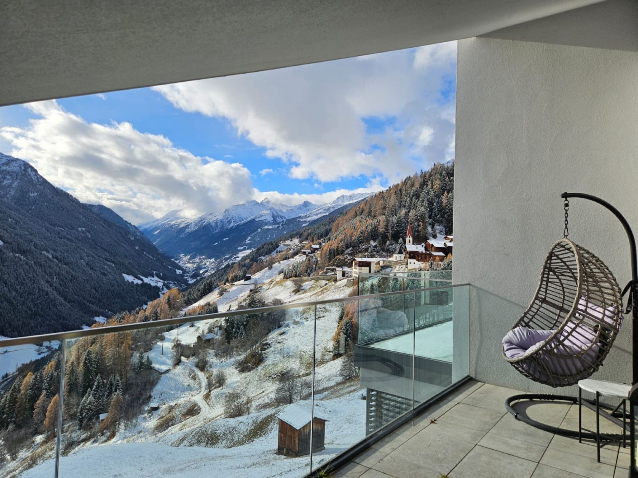 A balcony with a hanging chair, a glass balustrade, and a view of a snowy mountain landscape with buildings.