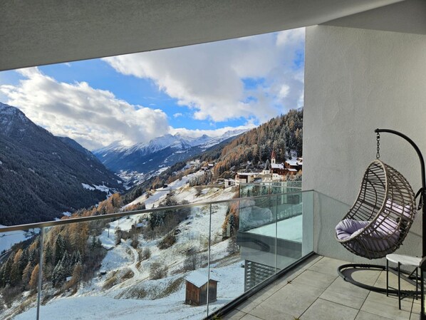 Un balcón con una silla colgante, barandilla de cristal y vista a un paisaje montañoso nevado con edificios.