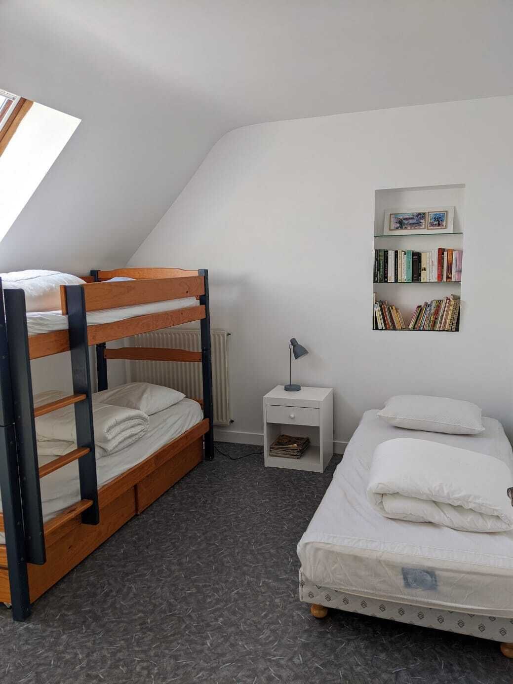 Una habitación tipo dormitorio con literas, una mesita de noche, una estantería y una ventana.