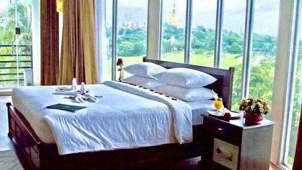 A neatly made bed with white linens, a bedside table with a lamp, and a view of a temple through large windows. - My Hotel Myanmar (Yangon)