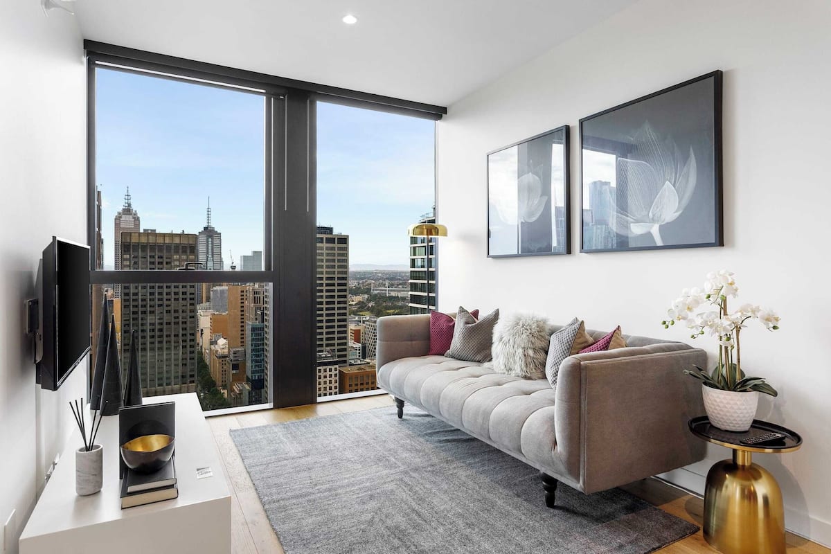 Un salón moderno con un sofá gris, un ventanal con vistas a la ciudad, un televisor de pantalla plana y dos obras de arte enmarcadas.