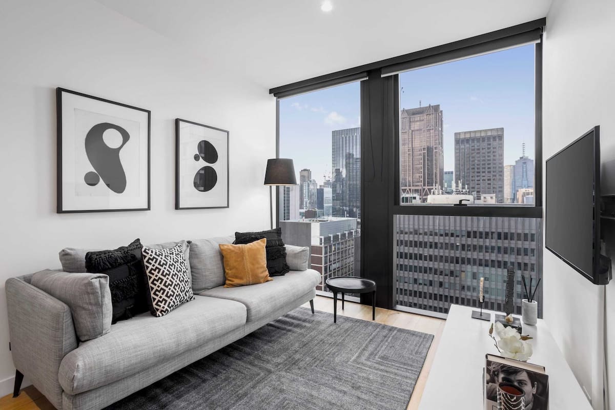 Un salón moderno con un sofá gris, cojines blancos y negros, una lámpara de pie negra y un amplio ventanal con vistas a la ciudad.