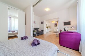 A modern hotel room with a large bed, a purple sofa, a dining area, and a kitchen in the background.