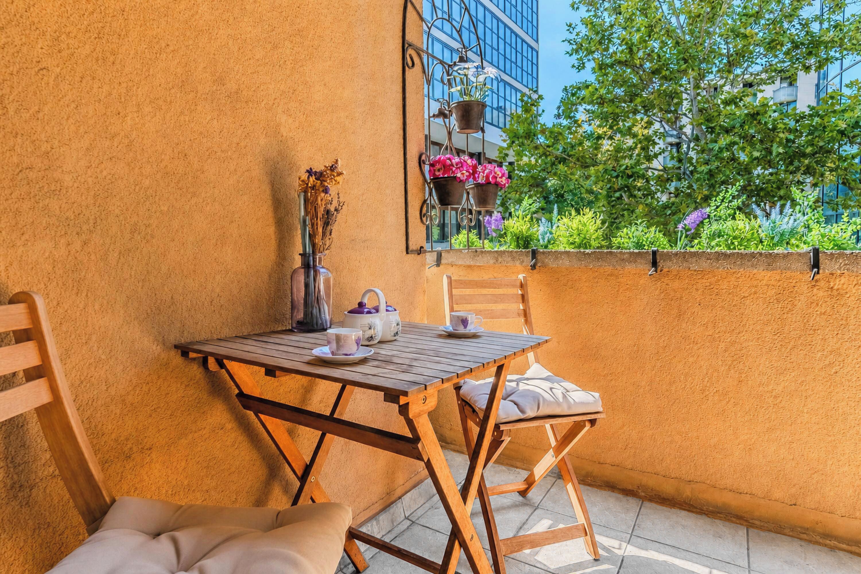 Une table en bois avec des chaises, un vase contenant des fleurs séchées et une théière sur un balcon d’où l’on peut admirer la verdure et les bâtiments.