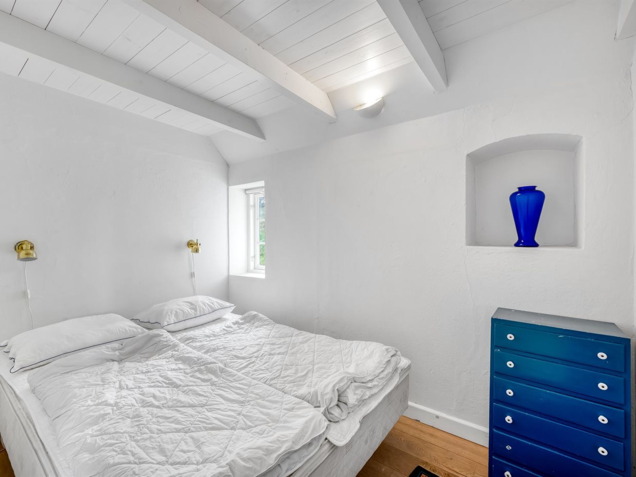 Una camera da letto con un letto, una cassettiera, una finestra e un vaso blu su uno scaffale.