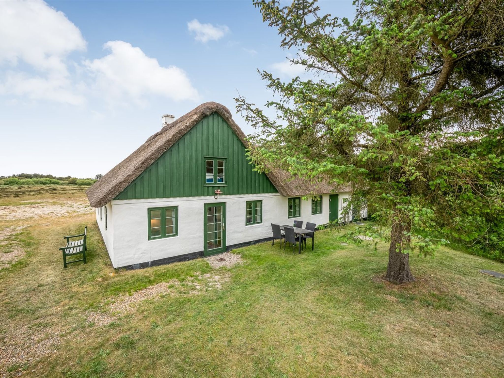 Une maison traditionnelle à toit de chaume, avec une façade verte et des murs blancs, un coin repas avec une table et des chaises, et un paysage verdoyant agrémenté d’un arbre.