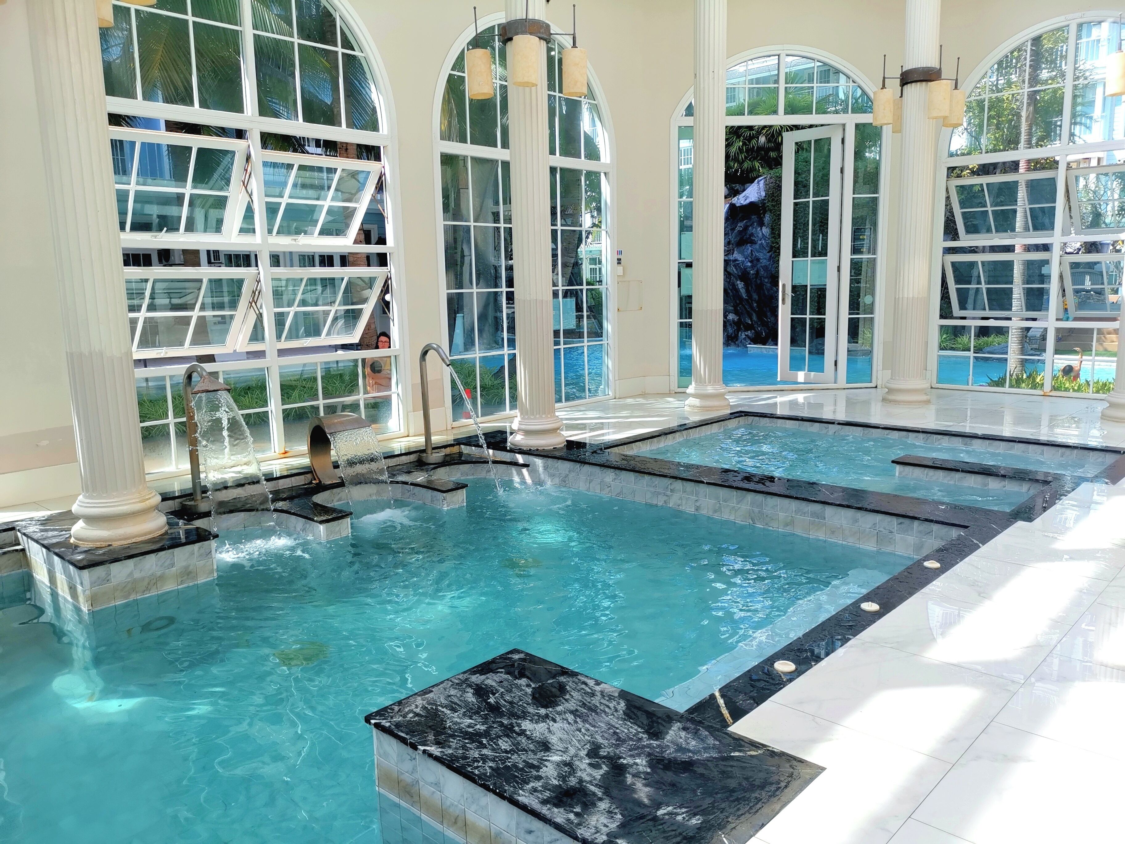 Ein großes Hallenbad mit mehreren Wasserspielen, umgeben von hohen Bogenfenstern und Säulen.