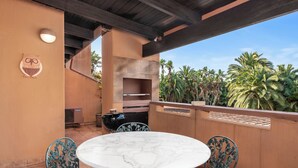 An outdoor patio with a round marble table and metal chairs, a built-in grill, and a view of palm trees.