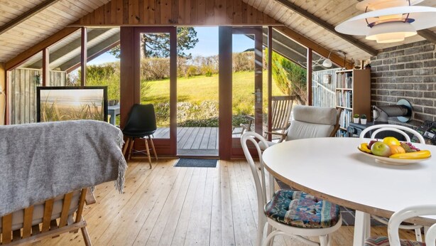 Een gezellig zolderkamer met houten balkenplafond, een eettafel met stoelen, een boekenkast en uitzicht op een groen landschap door grote ramen.