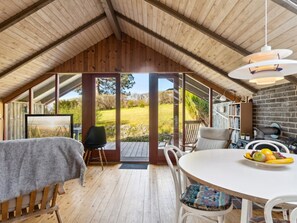 Ein gemütliches Dachzimmer mit Holzbalkendecke, einem Esstisch mit Stühlen, einem Bücherregal und Blick auf eine grüne Landschaft durch große Fenster.