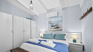 A bedroom with a bed, bedside tables, a bench, a painting of boats, and an air conditioning unit. - San Lameer Villa 2849 (Southbroom)