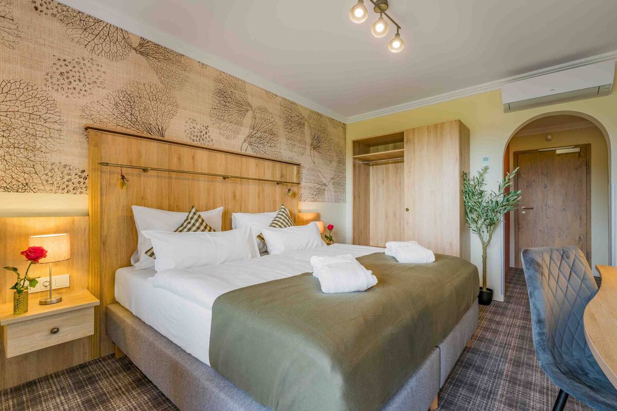A hotel room with a large bed, a wooden headboard, a nightstand with a lamp and a vase of flowers, a chair, and a potted plant.