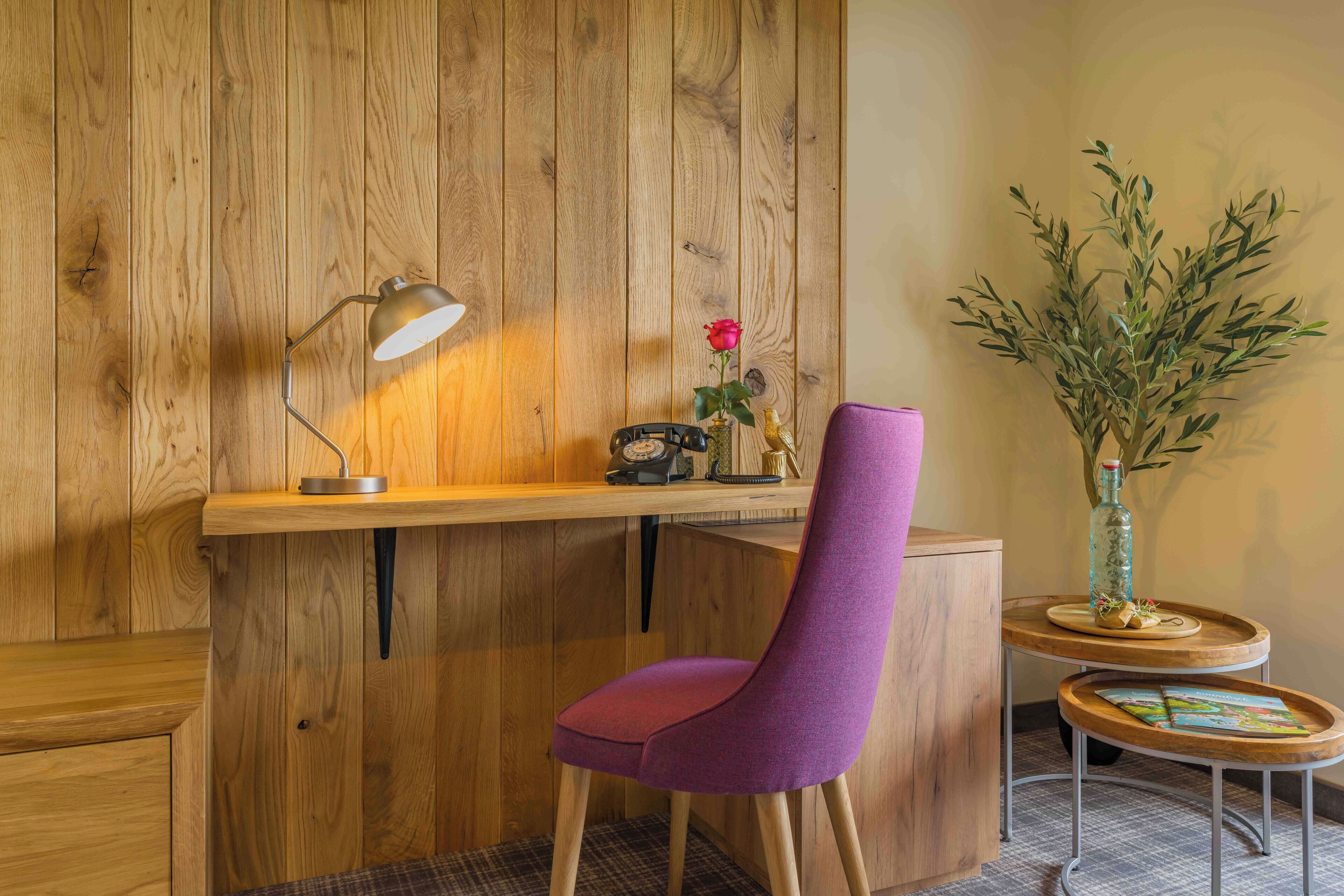 Un bureau en bois avec une lampe, une chaise, une petite table avec un vase et une plante, et un mur recouvert de panneaux de bois verticaux.