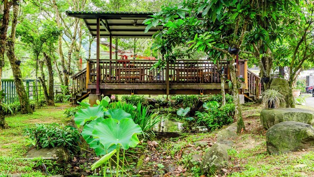 A wooden deck with a covered seating area surrounded by lush greenery and a small pond. - Green House (Puli)