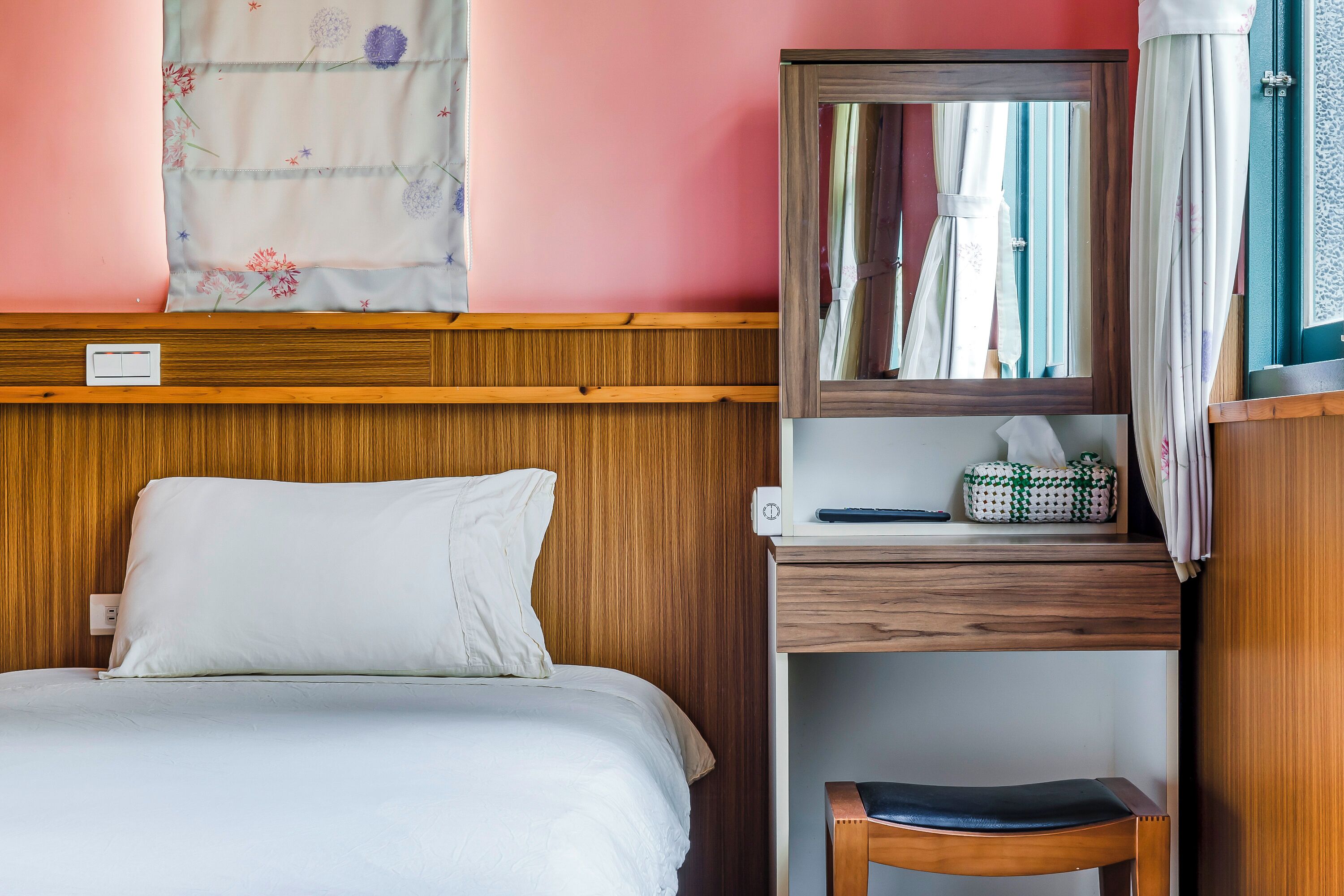 A neatly made bed with a white pillow and a wooden headboard. A small wooden desk with a mirror and a lamp is beside the bed.