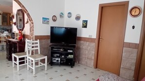 A living room with a TV, two white chairs, a sofa with a red cover, a wooden door, and a kitchen in the background. - Relax in Style:Spacious 4-Bedroom Villa Rosetta Near Palermo's Coastal Treasures (Carini)