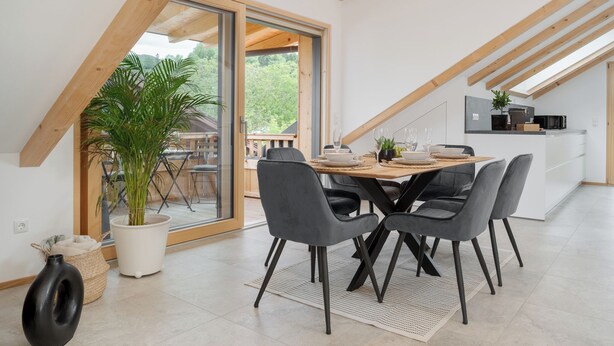 Ein Essbereich mit einem Holztisch, grauen Stühlen, einer Topfpflanze und Blick auf einen Balkon.