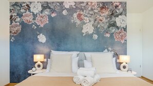 A neatly made bed with white linens and a rolled towel, flanked by two bedside tables with lamps, against a wall with a floral mural.
