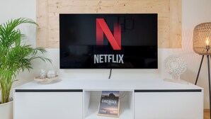 A modern living room with a large flat-screen TV mounted on a wooden wall, a white entertainment unit below, and a potted plant to the left.