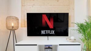 Ett modernt vardagsrum med en stor, platt-TV som är monterad på en vit vägg, en golvlampa och en krukväxt.