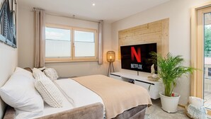 Ett modernt sovrum med en säng, en tv som är monterad på väggen, en krukväxt och ett fönster med persienner.