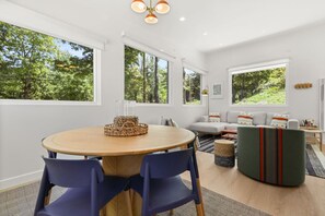 Un salón moderno con una mesa redonda de madera, sillas azules, un sofá verde y ventanas amplias con vistas a árboles.