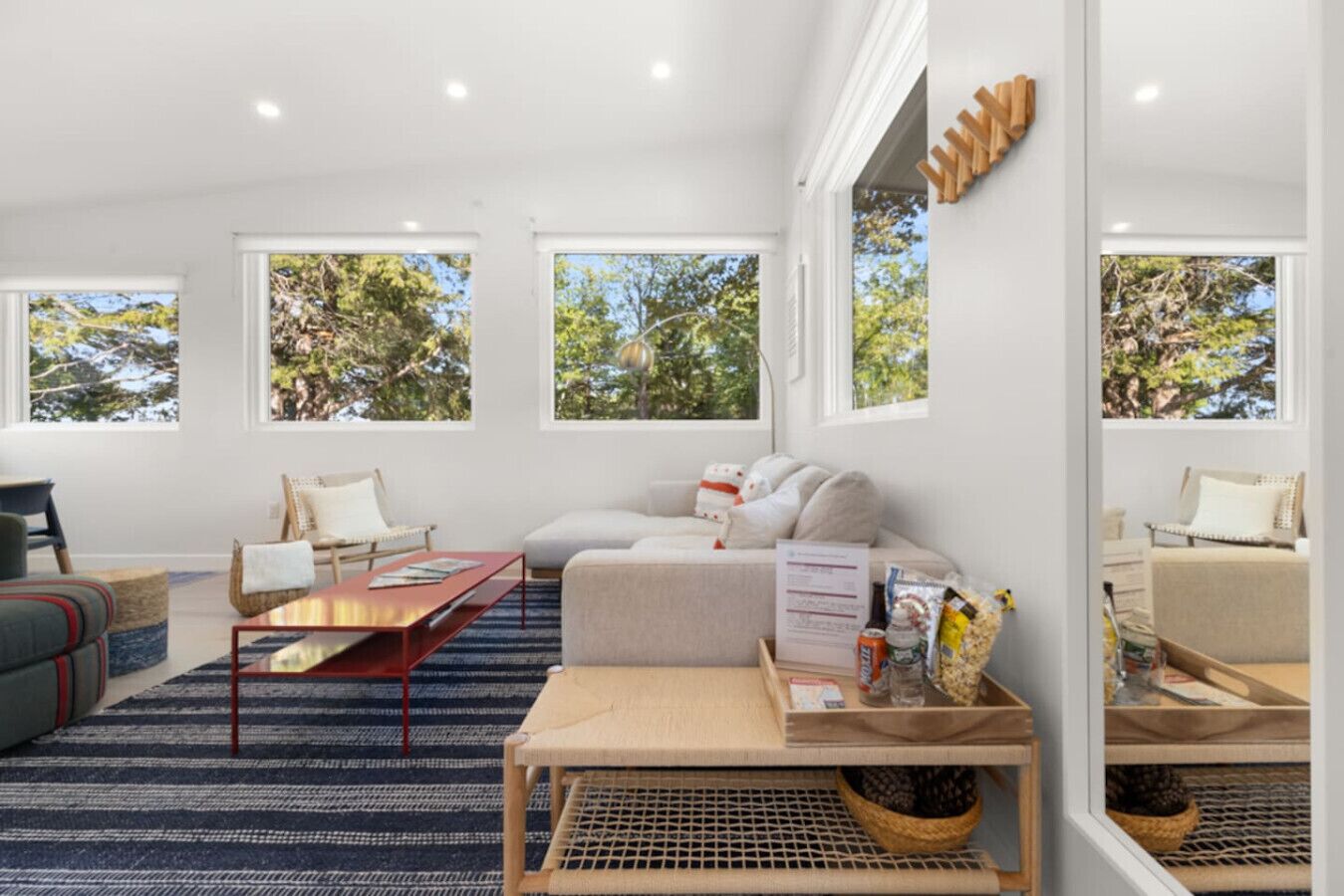 A modern living room with a sofa, coffee table, and chairs, large windows with a view of trees, and a wooden wall decor piece.