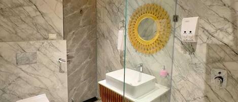 A bathroom with a marble wall, a glass-enclosed sink, a toilet, and a shower area.