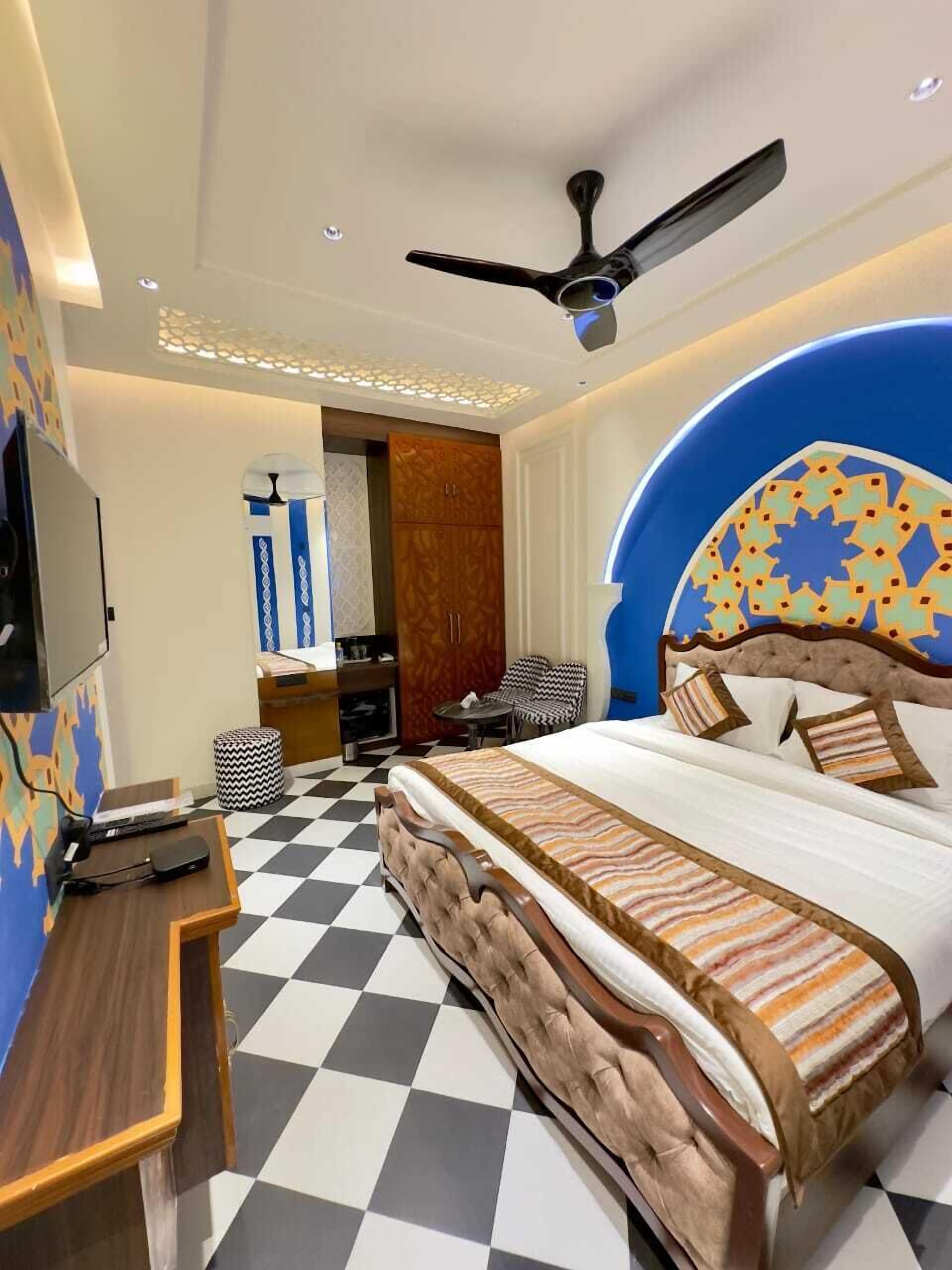 A hotel room with a checkered floor, a bed with a patterned headboard, a desk with a computer, a ceiling fan, and a TV mounted on the wall.