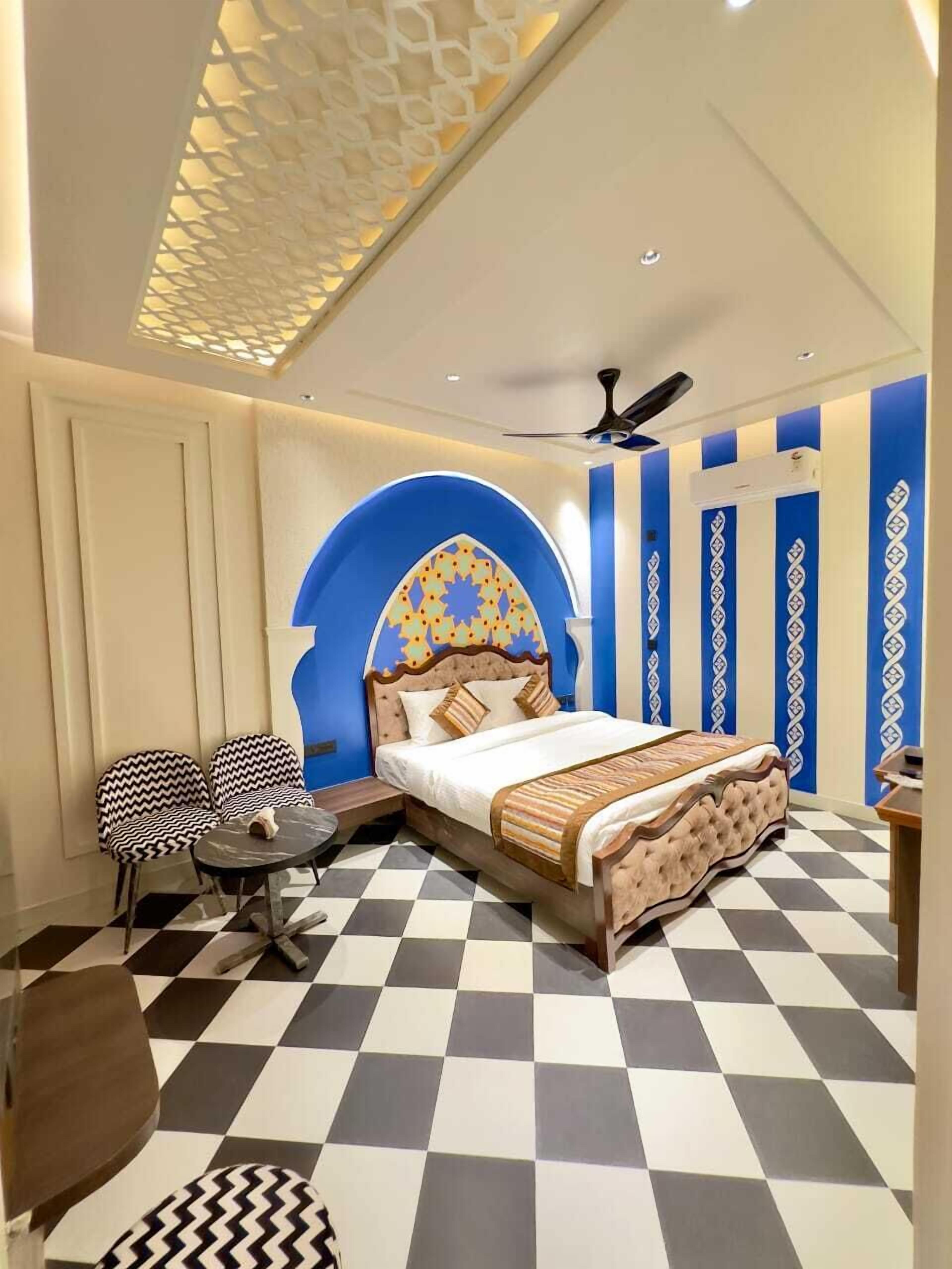A bedroom with a checkered floor, a bed with a patterned headboard, two chairs, a small table, and a ceiling fan.
