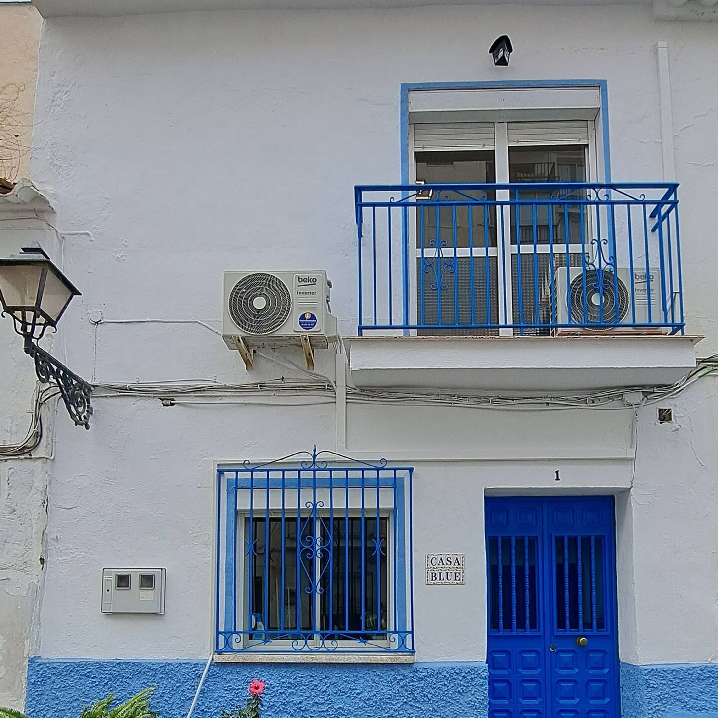 Un edificio de dos pisos con puerta y ventanas azules, un balcón con barandal azul y dos unidades de aire acondicionado montadas en la pared.