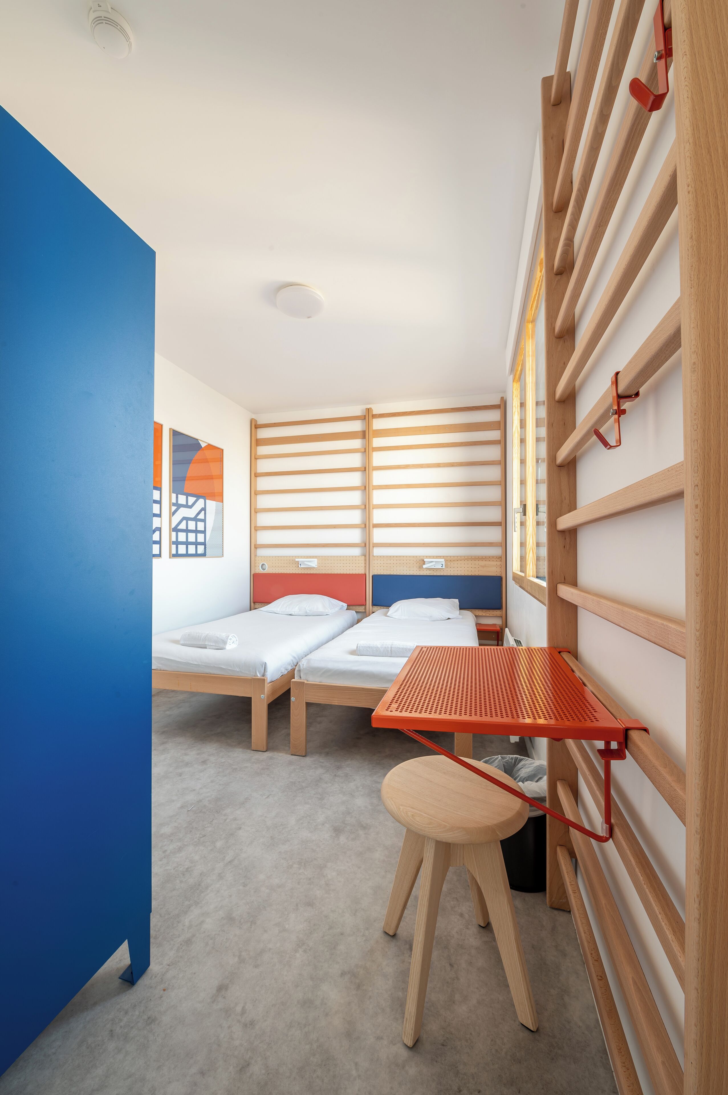 Een compacte kamer met twee bedden, een klein krukje en een rode plank.