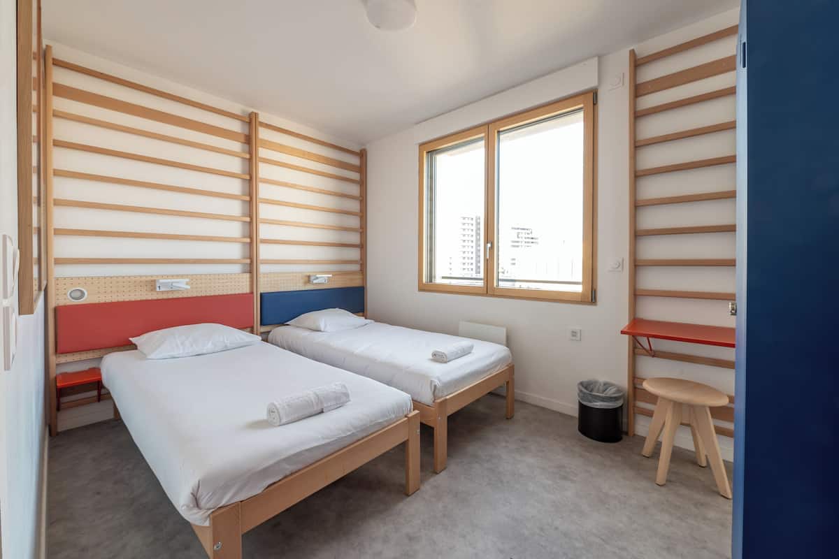 Ein Zimmer mit zwei Betten, einem Fenster, einer roten Bank, einem Hocker und einem Mülleimer.