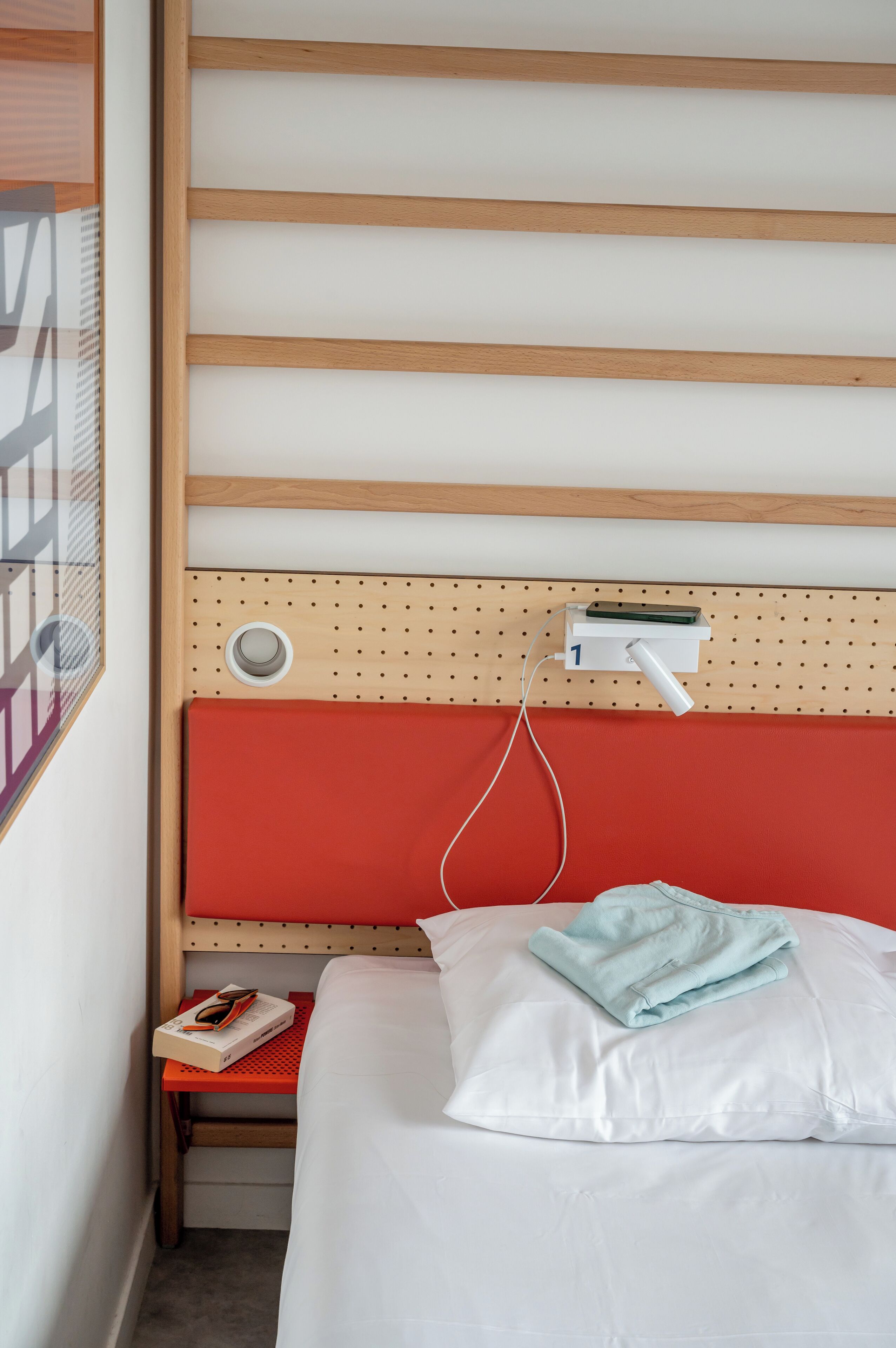 Habitación de hotel con cabecera de madera, pared de acento roja, cama blanca con toalla azul y mesita de noche con un libro y un control remoto.