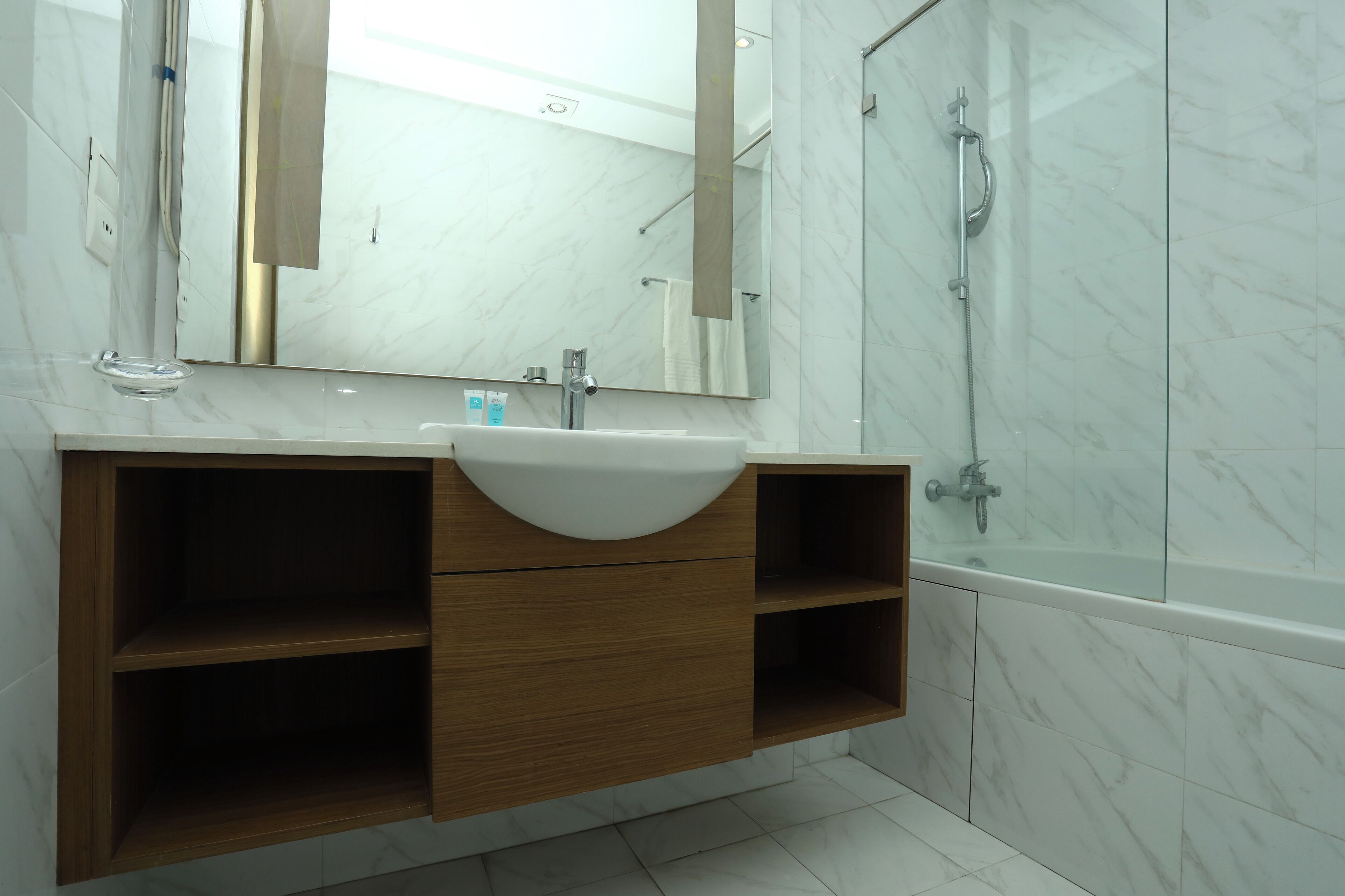 A bathroom with a marble-tiled wall, a wooden vanity with a sink, a mirror, and a glass-enclosed shower.