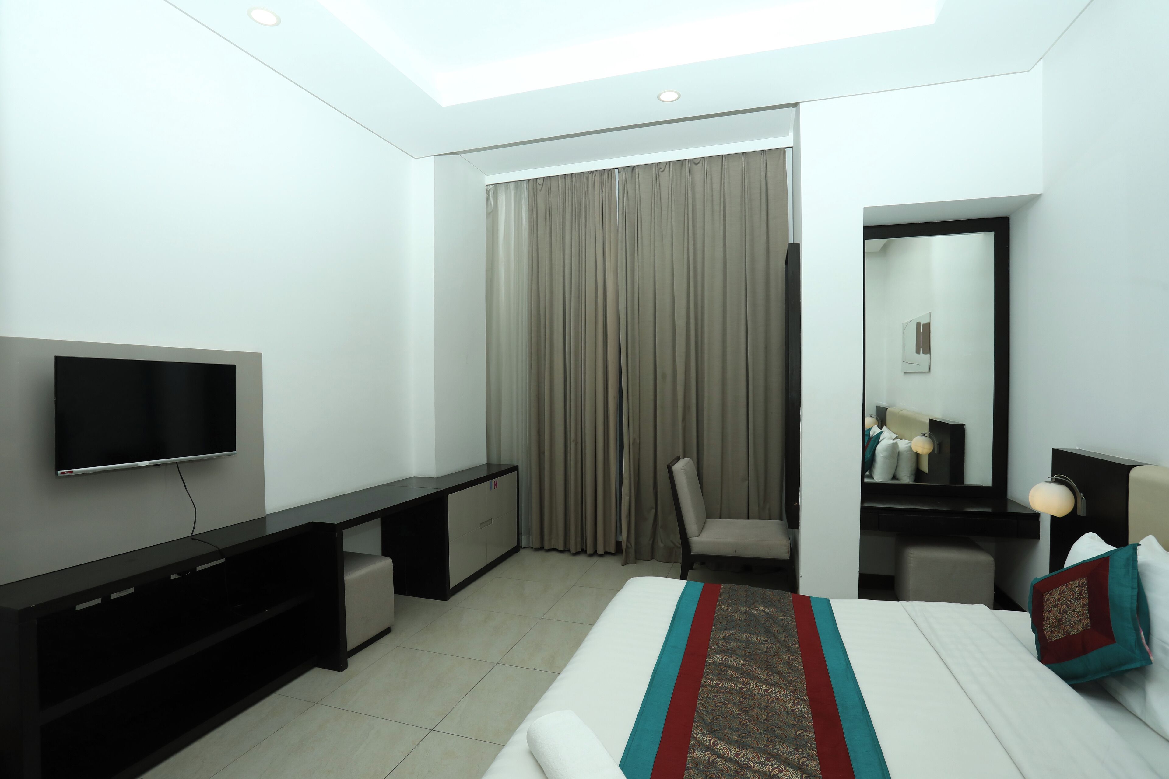 Una habitación de hotel moderna con una televisión de pantalla plana, un escritorio, una silla, una cama con cubrecama estampado y un ventanal con cortinas.