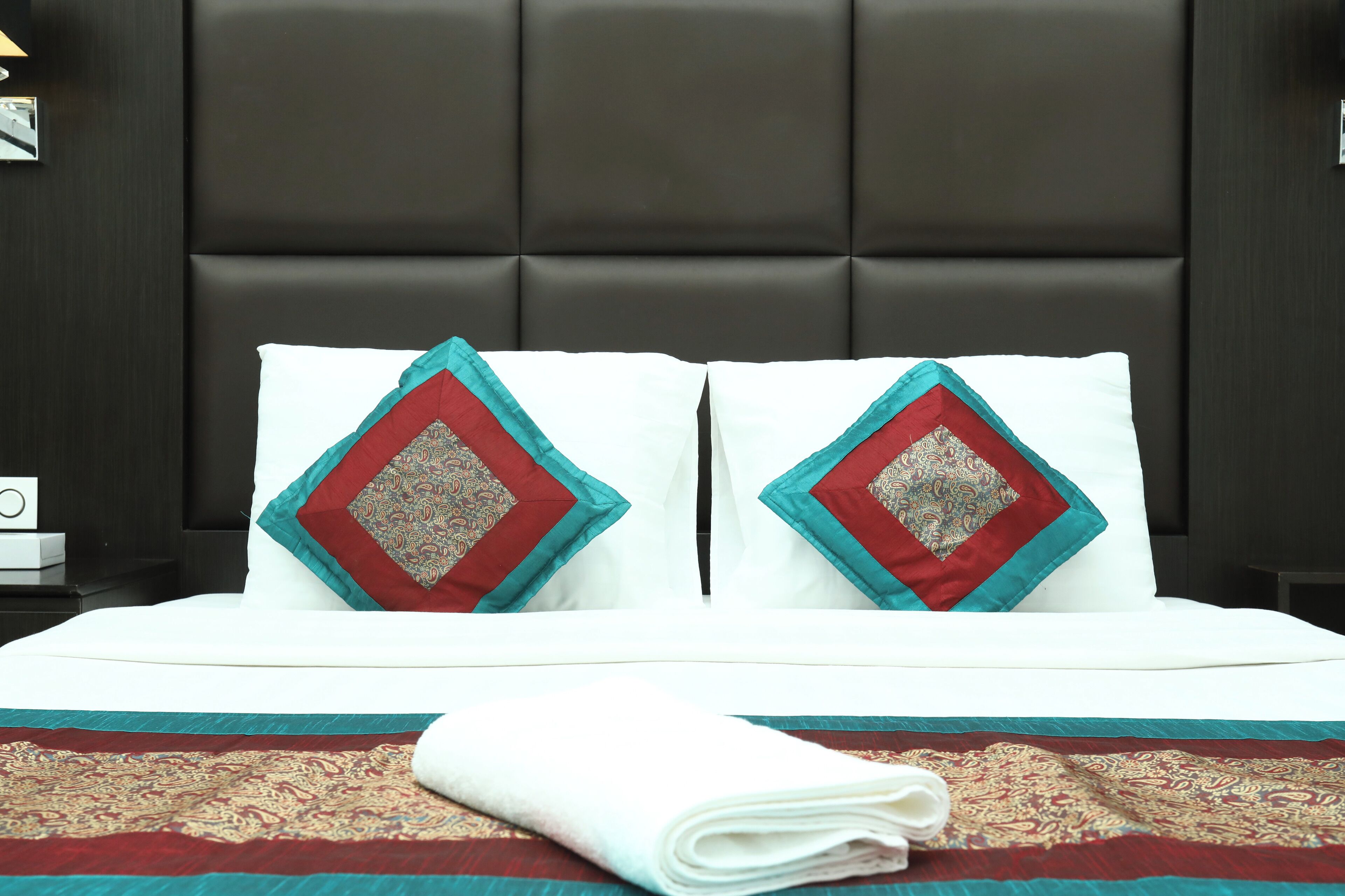 A neatly made bed with a patterned bedspread, two pillows with a geometric design, and a folded white towel on the bed.