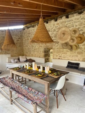 A rustic dining area with a wooden table set for a meal, a woven bench, and a stone wall with woven baskets.