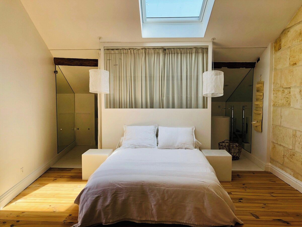 A bedroom with a skylight, a bed with white bedding, a bedside table, and a large mirror.